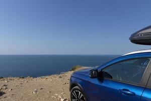 un coche azul con una baca en el borde de un acantilado contra el cielo azul y el mar. concepto de viaje en coche. foto