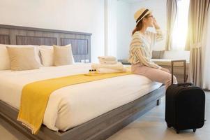 mujer turista sentada en la cama y mirando la hermosa vista fuera de la habitación con su equipaje en la habitación del hotel después del check-in. conceptual de viaje y alojamiento. foto