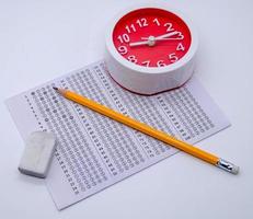 Answers sheet with yellow sharp pencil, clock and rubber isolated on white background. Top view of them. Take the exam timely concept. photo