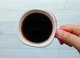 el primer plano y la vista superior de una mano sostienen una taza roja de café negro caliente con fondo borroso por la mañana. concepto de comienzo del día. foto