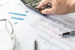 financial accountant, work from home, closeup man's hand touching calculator button on desktop, check account number on paper and calculator, large numbers, graph, glasses on paper, blurry background photo