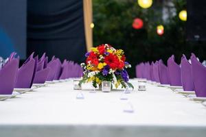 El ramo de flores rojas, amarillas, moradas y blancas está colocado en la larga mesa de cubierta blanca y listo para una cena de lujo en el campo del jardín. foto