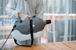 Professional technical man in prevention suit sprays sterilizing solutionby electrical spray machine in the indoor glass house room. photo