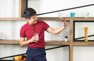 Smart, young and healthy Asian man holding a glass of milk. Young man using smartphone in the loft style kitchen room to selfie. photo