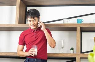 Smart, young and healthy Asian man holding a glass of milk. Young man using smartphone in the loft style kitchen room photo