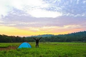 The man who stand next to the tent in the meadow put his hand up  with wide sky and cloud photo