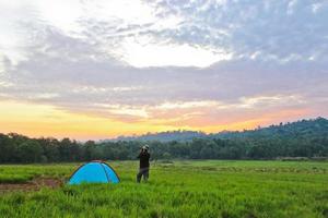 The man who stand next to the tent in the meadow were taking the landscape photograph in front of him photo