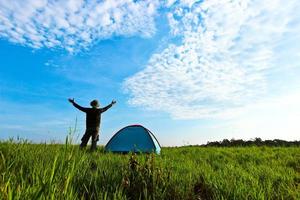 The man who stand next to the tent put his hand up in the meadow with wide blue sky and white cloud photo