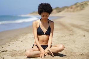 Positive black girl in bikini sitting on the sand of a beach gazing thoughtfully at the sand. photo