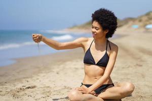 Positive black woman in bikini sitting playing with sand on the beach. photo