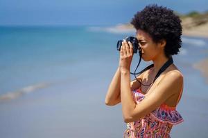 Young black woman with afro hair in summer dress, taking pictures with an SLR camera on the beach. photo