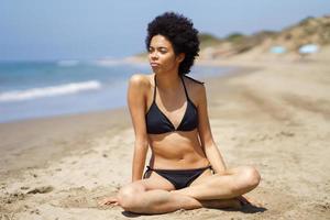 Relaxed black woman in bikini sitting on the sand of a tropical beach looking out to sea. photo