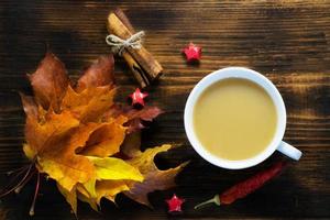 Mug of hot coffee with milk and spices and autumn leaves on wooden table. Morning cup of cappuccino coffee with cinnamon and maple leaves. photo