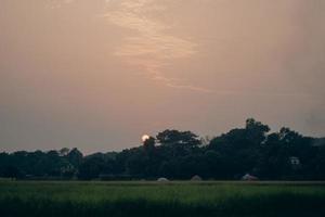 Red sun hiding behind the tree, sunrise view on a rural area. Sunset view before dusk on greenfield. A beautiful sunset or sunrise photoshoot with a cloudy sky and red horizon. photo