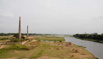 Brick kilns on a greenfield beside Titas river with a bunch of haystack photoshoots. Greenfield and tree horizon with two brick kilns. Beautiful river view with green fields and cloudy sky. photo