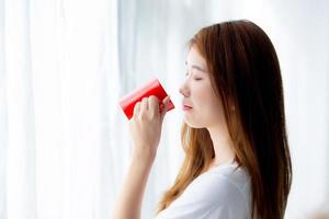 Beautiful of portrait young asian woman with drink acup of coffee standing curtain window background in bedroom, girl relax in morning at home, lifestyle concept. photo