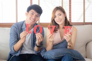 Beautiful young asian couple sitting on sofa holding word love together in living room, family husband and wife smiling with happy, man and woman lover wearing casual with relax, lifestyle concept. photo