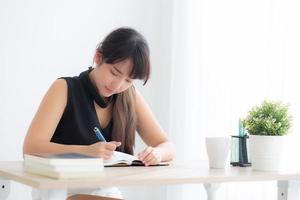 Beautiful young asian woman smiling sitting study and learning writing notebook and diary in the living room at home, girl homework, business woman working on table, education concept. photo