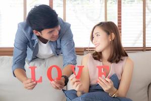Beautiful young asian couple sitting on sofa holding word love together in living room, family husband and wife smiling with happy, man and woman lover wearing casual with relax, lifestyle concept. photo