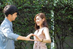 Beautiful happy young couple fun making gesture heart shape with hand outdoor together, man and woman with relation feeling love with symbol and sign in the garden, lover and romantic concept. photo