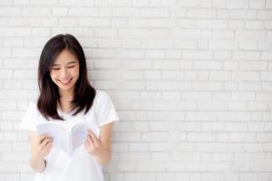 Beautiful of portrait young asian woman happiness relax standing reading book on concrete cement white background at home, girl happy study content literature, education and lifestyle concept. photo