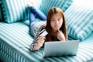 Beautiful of portrait young asian woman lying think use credit card with laptop, Content girl shopping online and payment with notebook computer on sofa, lifestyle concept. photo