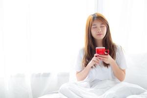 hermoso retrato de una joven asiática con una taza de café de pie en el fondo de la ventana de la cortina en el dormitorio, la chica se relaja por la mañana en casa, el concepto de estilo de vida. foto