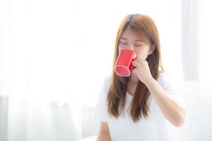 hermoso retrato de una joven asiática con una taza de café de pie en el fondo de la ventana de la cortina en el dormitorio, la chica se relaja por la mañana en casa, el concepto de estilo de vida. foto