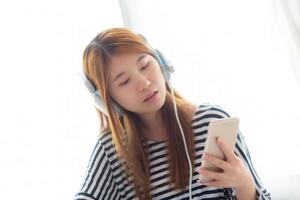 hermosa joven asiática disfruta escuchando música con auriculares y sosteniendo un teléfono móvil inteligente mientras está sentada en el dormitorio, relaja a la chica con el concepto de auriculares, ocio y tecnología. foto