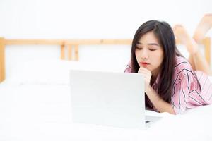 hermosa mujer joven asiática acostada en la cama usando una computadora portátil en el dormitorio para el ocio y relajarse, independiente con un cuaderno de trabajo para niñas, concepto de comunicación. foto