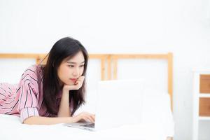 hermosa mujer joven asiática acostada en la cama usando una computadora portátil en el dormitorio para el ocio y relajarse, independiente con un cuaderno de trabajo para niñas, concepto de comunicación. foto