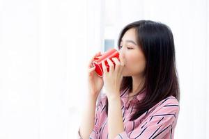 Beautiful of portrait young asian woman with drink a cup of coffee standing curtain window background in bedroom, girl relax in morning at home, lifestyle concept. photo