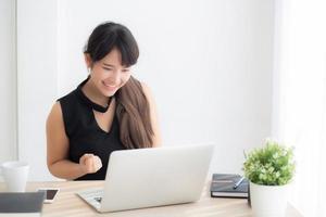 hermoso retrato mujer joven asiática sonríe trabajando en una computadora portátil en línea con feliz satisfecha sentada en el escritorio de la oficina, chica usando un cuaderno con el concepto emocionado y alegre, de negocios y de éxito. foto