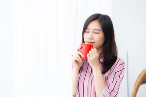 hermoso retrato de una joven asiática con una taza de café de pie en el fondo de la ventana de la cortina en el dormitorio, la chica se relaja por la mañana en casa, el concepto de estilo de vida. foto