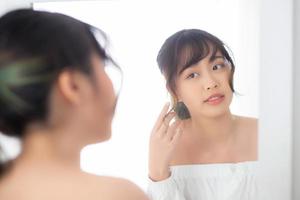 Beauty portrait young asian woman smiling look at mirror of checking skin care caucasian with wellness in the bedroom, beautiful girl happy touching face in reflection for health, lifestyle concept. photo