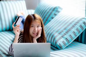 Beautiful of portrait young asian woman lying users credit card with laptop, Content girl shopping online and payment with notebook computer on sofa, lifestyle concept. photo