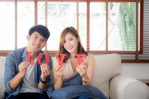 Beautiful young asian couple sitting on sofa holding word love together in living room, family husband and wife smiling with happy, man and woman lover wearing casual with relax, lifestyle concept. photo