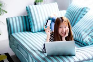 Beautiful of portrait young asian woman lying users credit card with laptop, Content girl shopping online and payment with notebook computer on sofa, lifestyle concept. photo