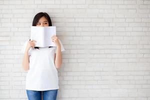 hermoso retrato joven mujer asiática feliz escondida detrás de abrir el libro con cemento o fondo de hormigón, niña de pie leyendo para el concepto de aprendizaje, educación y conocimiento. foto