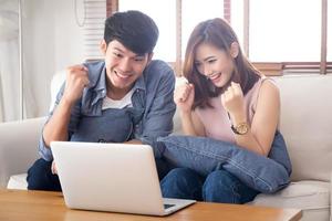 Beautiful portrait asian young couple working laptop with smile and happy sitting on couch at living room, man and woman on sofa using notebook computer with excited, business and success concept. photo