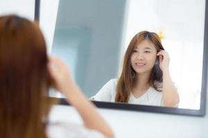 hermosa de retrato mujer joven asiática examinando con cara y sonrisa mirando en el espejo en el dormitorio, chica belleza de maquillaje y vestida con reflexión, estilo de vida y concepto de bienestar. foto