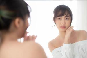 Beauty portrait young asian woman smiling look at mirror of checking skin care caucasian with wellness in the bedroom, beautiful girl happy touching face in reflection for health, lifestyle concept. photo