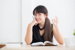 hermoso retrato joven mujer asiática sonriendo sentada estudiando y aprendiendo a escribir cuaderno y diario en la sala de estar en casa, tarea de niña, mujer de negocios trabajando en la mesa, concepto de educación. foto