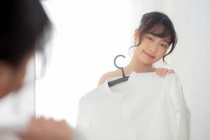 joven mujer asiática sonriendo con ropa tratando de vestirse con un espejo de reflexión moderno y de aspecto en la habitación, la chica elige ropa de moda con un concepto de estilo de vida feliz y alegre. foto