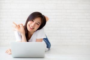 Beautiful portrait asian young woman working online laptop with excited lying on floor brick cement background, freelance girl smile using notebook computer with happy, business and lifestyle concept. photo