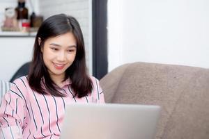 Beautiful of portrait asian young woman working online laptop with smile and happy sitting on sofa at living room, girl using notebook computer with connect to internet, business and success concept. photo