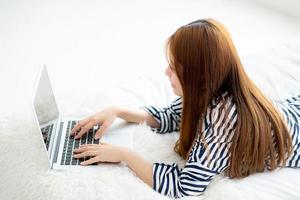Beautiful asian young woman lying on bed using laptop at bedroom for leisure and relax, freelance with girl working notebook, communication concept. photo