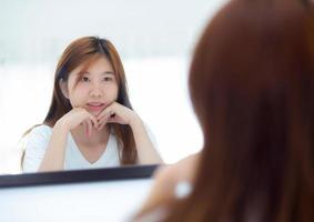 hermosa de retrato mujer joven asiática examinando con cara y sonrisa mirando en el espejo en el dormitorio, chica belleza de maquillaje y vestida con reflexión, estilo de vida y concepto de bienestar. foto