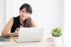 Beautiful young asian woman smiling working and typing on laptop computer at desk office with professional, girl using notebook checking email or social network, business and lifestyle concept. photo