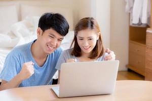 Beautiful portrait young asian couple working laptop with smile and happy sitting in bedroom, man and woman using notebook computer with excited and satisfied, business and success concept. photo